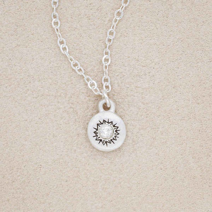 Dainty Sunburst Necklace cast in Sterling Silver and set with a 3mm cubic zirconia stone, on a beige background