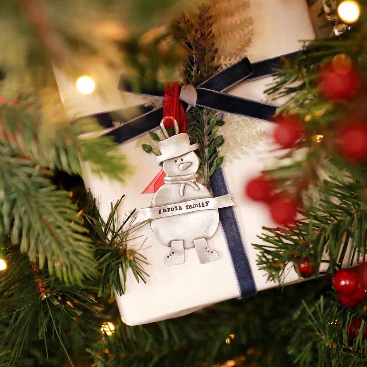 Snowman ornament hand-molded and cast in pewter hung from a sheer red ribbon and personalized with a phrase being placed on a gift box