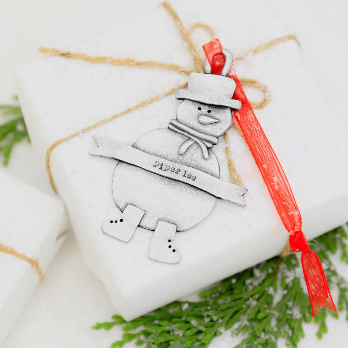 Snowman ornament hand-molded and cast in pewter hung from a sheer red ribbon and personalized with a phrase being placed on a Christmas tree