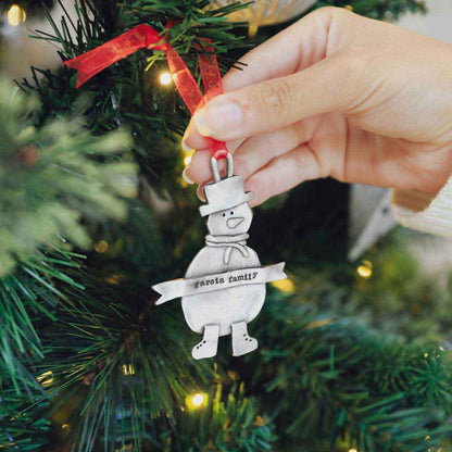 Snowman ornament hand-molded and cast in pewter hung from a sheer red ribbon and personalized with a phrase being placed on a Christmas tree